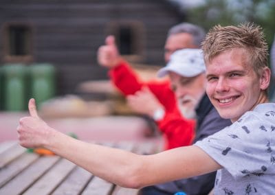 3 jongens/mannen zitten aan een picknicktafel. 2 met de duim onhoog en lachtend gezicht